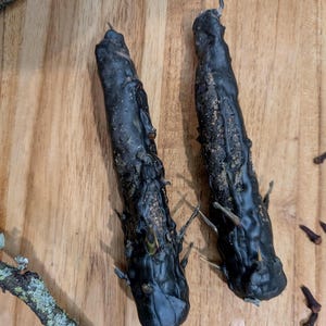 Puede incluir: Dos velas negras texturizadas con púas sobresalientes se exhiben sobre una superficie de madera. Las velas tienen un aspecto rústico, con imperfecciones visibles y un aspecto ligeramente derretido. El fondo incluye ramitas y clavos.