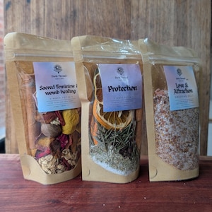 Puede incluir: Tres bolsas verticales con ventanas transparentes que contienen mezclas botánicas. La bolsa izquierda está etiquetada Sacred feminine womb healing, la central Protection y la derecha Love & Attraction, todas de Dark Thread.