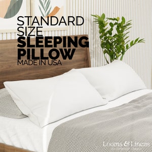 May include: A standard size white sleeping pillow on a bed with a gray and white checkered bedspread. The headboard is made of wood and has the text "STANDARD SIZE SLEEPING PILLOW MADE IN USA" printed on it.