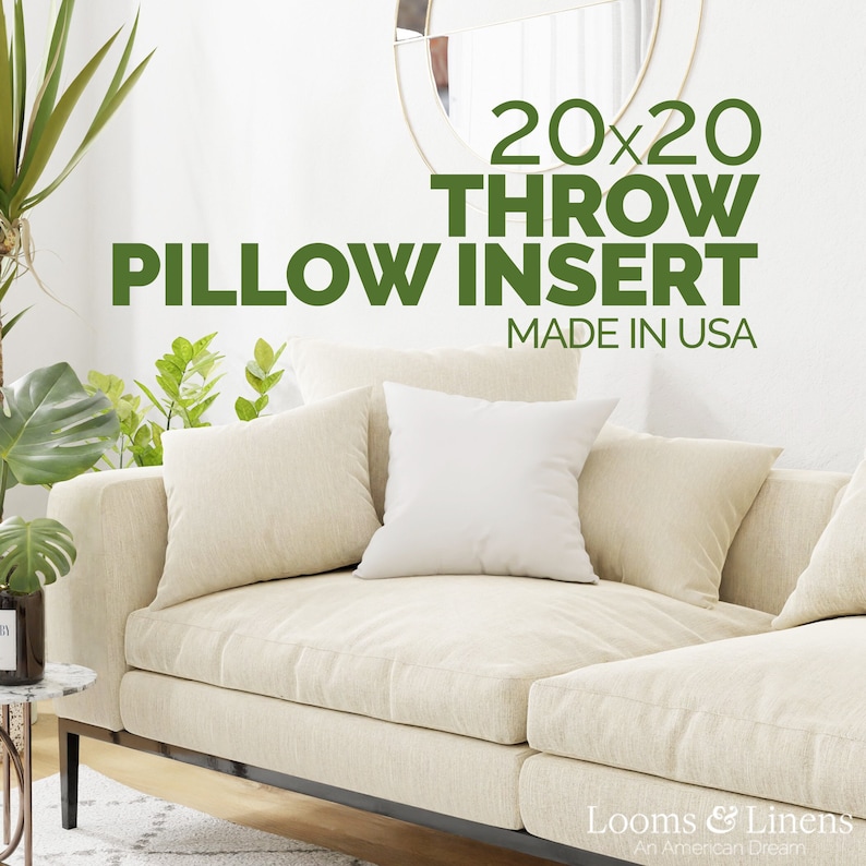 May include: A white couch with two throw pillows. The couch is in a living room with plants and a side table. The text on the wall reads "20x20 Throw Pillow Insert Made in USA".