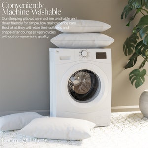 May include: White washing machine with a white door and a silver control panel. Two white pillows are stacked on top of the washing machine. The washing machine is sitting on a white tiled floor with a geometric pattern. The text "Conveniently Machine Washable" is visible in the top left corner of the image.