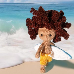 Può includere: Bambola all'uncinetto di un surfista con capelli ricci marroni, che indossa pantaloncini gialli e tiene una tavola da surf bianca e blu. La bambola è su una spiaggia sabbiosa con l'oceano sullo sfondo.
