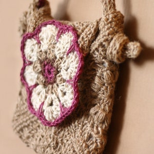 Könnte beinhalten: Eine kleine, gehäkelte Tasche mit einem braunen und weißen Blumendesign. Die Blume hat einen rosa Rand und ist an der Vorderseite der Tasche befestigt.