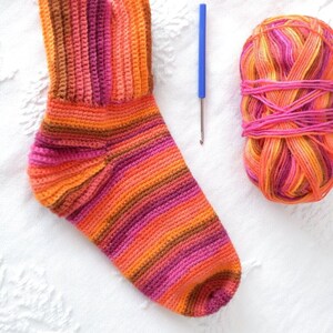 Può includere: Un calzino lavorato a uncinetto in tonalità rosa, arancio e marrone. Il calzino è appoggiato su una superficie bianca accanto a un gomitolo di lana degli stessi colori e un uncinetto.