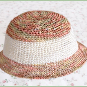 Può includere: Un cappello da sole all'uncinetto con un'ampia tesa. Il cappello è realizzato con filato multicolore in tonalità di rosa, verde e beige. Il cappello ha una fascia bianca attorno alla corona.