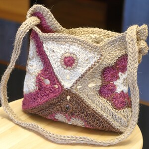 Könnte beinhalten: Eine gehäkelte Tasche mit einem braunen, weißen und rosafarbenen Blumenmuster. Die Tasche hat einen langen Riemen und ist aus Naturfasern gefertigt.
