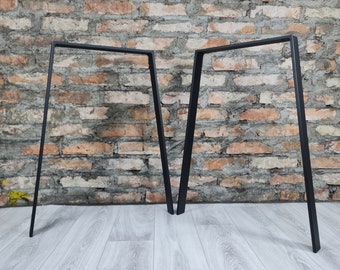 Op de afbeelding: Twee zwarte metalen A-vormige tafelpoten, staand op een lichte houten vloer voor een bakstenen muur.