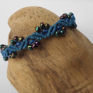 Könnte beinhalten: Ein blaues Makramee-Armband mit schillernden blauen und schwarzen Perlen. Das Armband liegt auf einer Holzoberfläche.