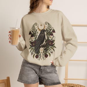 Peut inclure: Un sweat-shirt à col rond beige orné d'un motif central représentant un corbeau noir perché sur une branche, entouré de fleurs rouge foncé, de feuilles de fougère vertes et d'un croissant de lune doré avec une étoile, porté avec un short en jean gris.