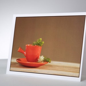 May include: A greeting card featuring a vibrant red teacup and saucer, adorned with sprigs of greenery. The teacup has a distinctive handle and sits on a matching saucer. The background is a warm brown, and the card is displayed on a light-colored surface.