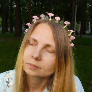 May include: A woman with long blonde hair wearing a crown of red and white mushrooms. She is wearing a light blue shirt with a white crane pattern.