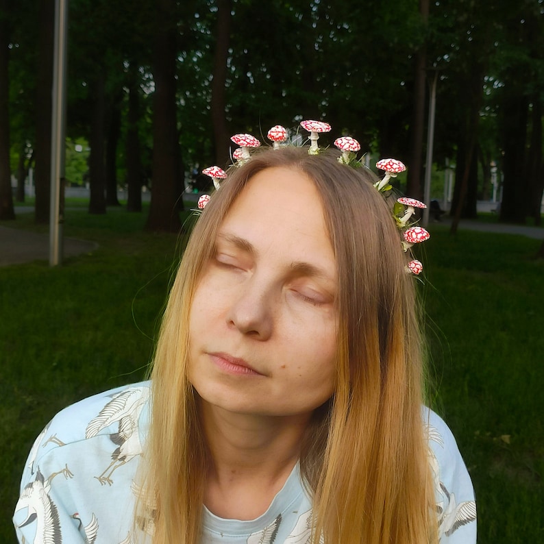 May include: A woman with long blonde hair wearing a crown of red and white mushrooms. She is wearing a light blue shirt with a white crane pattern.