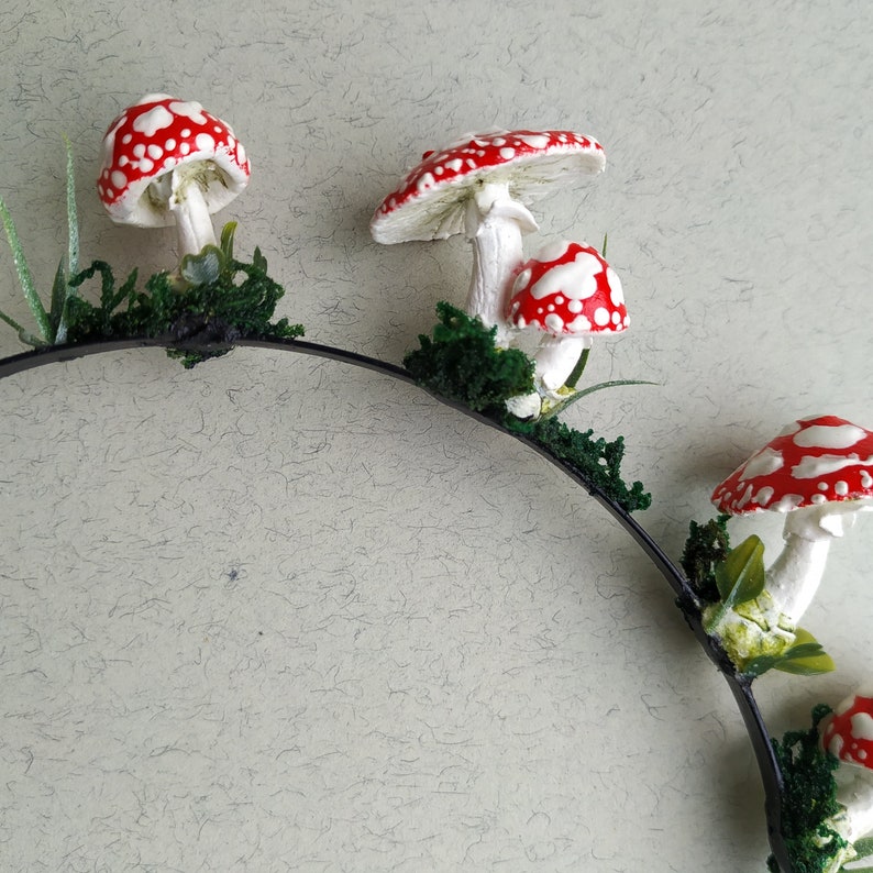 May include: A black headband with red and white mushroom decorations. The mushrooms are attached to the headband with green foliage.
