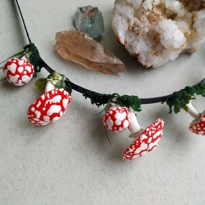 May include: A black headband with red and white mushroom charms and green foliage accents. The headband is decorated with small, realistic-looking mushrooms.