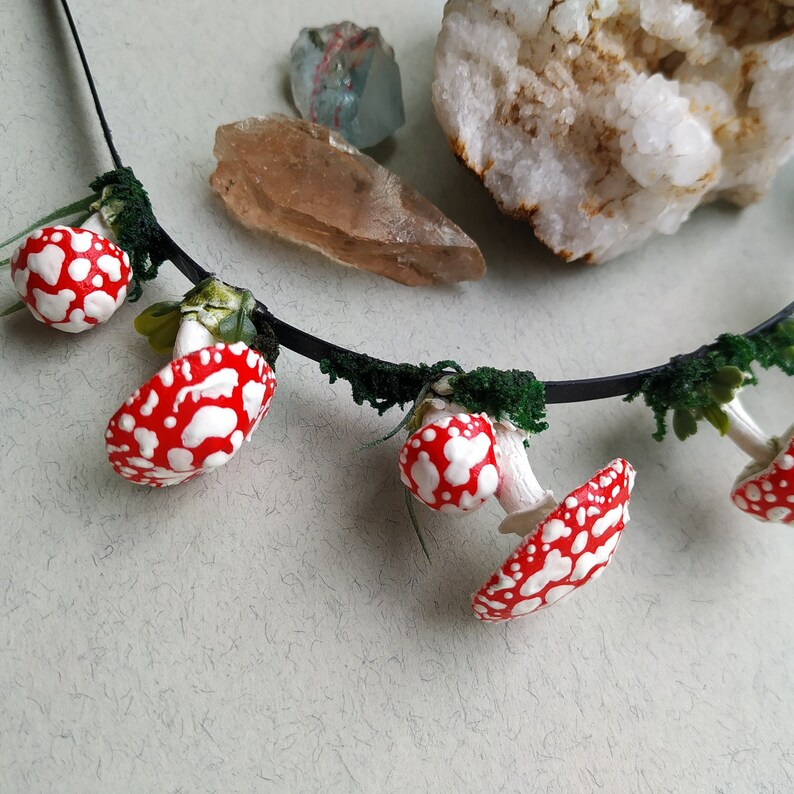 May include: A black headband with red and white mushroom charms and green foliage accents. The headband is decorated with small, realistic-looking mushrooms.