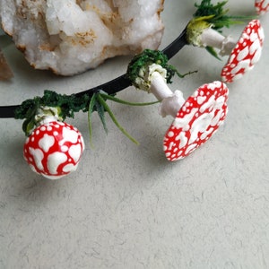May include: A black headband with red and white mushroom accents. The mushrooms are attached to the headband with green foliage.