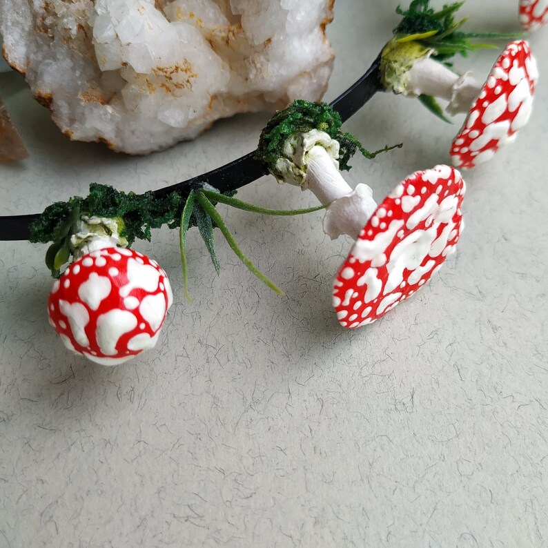 May include: A black headband with red and white mushroom accents. The mushrooms are attached to the headband with green foliage.