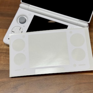 Pu&ograve; includere: Una console di gioco portatile Nintendo 3DS bianca con una skin protettiva bianca applicata. La skin ha ritagli per i pulsanti e lo schermo. La console &egrave; su una superficie di legno.