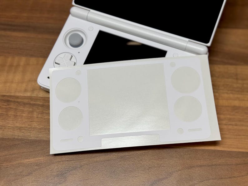 Pu&ograve; includere: Una console di gioco portatile Nintendo 3DS bianca con una skin protettiva bianca applicata. La skin ha ritagli per i pulsanti e lo schermo. La console &egrave; su una superficie di legno.