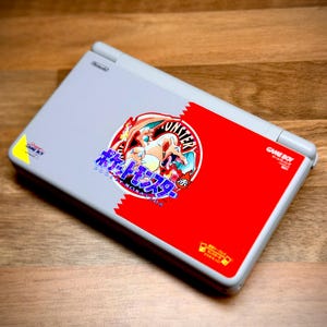 May include: A gray Nintendo Game Boy Advance SP console with a red and white design featuring a Pok&eacute;mon graphic. The console is closed and rests on a wooden surface.