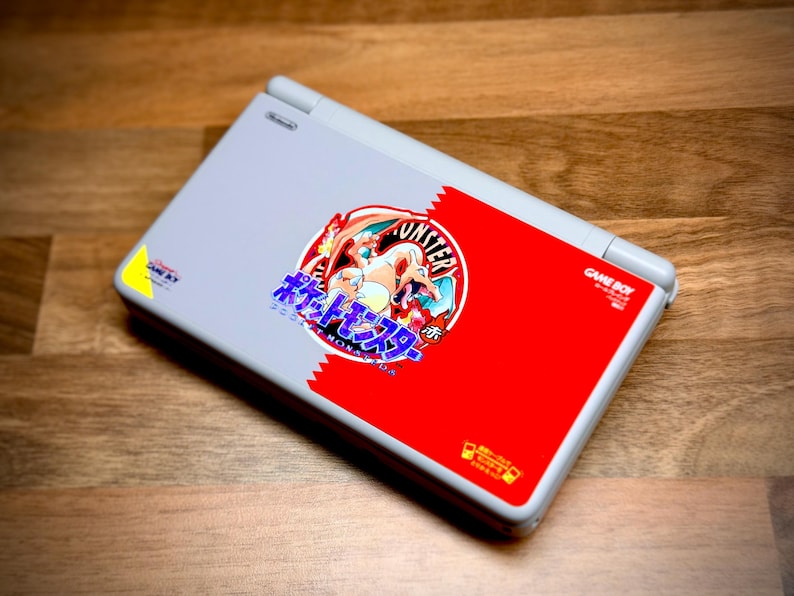 May include: A gray Nintendo Game Boy Advance SP console with a red and white design featuring a Pok&eacute;mon graphic. The console is closed and rests on a wooden surface.