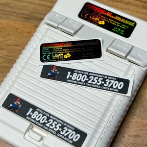 K&ouml;nnte beinhalten: Eine wei&szlig;e Nintendo Game & Watch: Super Mario Bros. Handheld-Konsole mit einem schwarz-wei&szlig;en Aufkleber mit der Aufschrift "INSTALLATION, MAINTENANCE OR SERVICE 1-800-255-3700 NINTENDO WORLD CLASS SERVICE, CENTER HOTLINE".