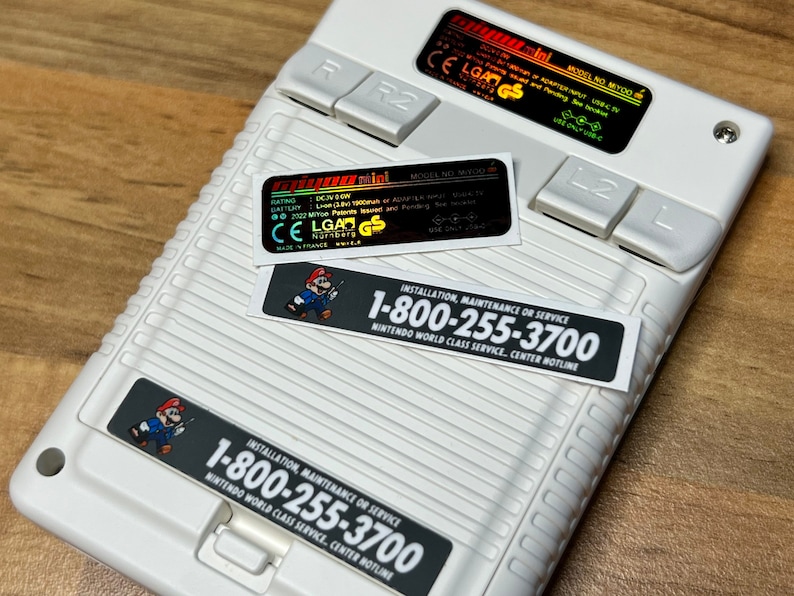 K&ouml;nnte beinhalten: Eine wei&szlig;e Nintendo Game & Watch: Super Mario Bros. Handheld-Konsole mit einem schwarz-wei&szlig;en Aufkleber mit der Aufschrift "INSTALLATION, MAINTENANCE OR SERVICE 1-800-255-3700 NINTENDO WORLD CLASS SERVICE, CENTER HOTLINE".