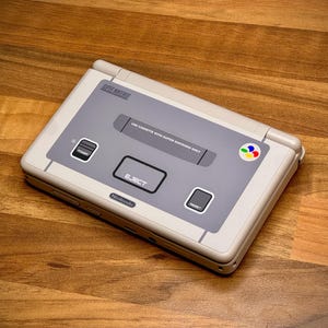 May include: A handheld Super Nintendo Entertainment System (SNES) in light grey and off-white. It has a grey central panel with an 'EJECT' button and a multi-coloured logo. The console is on a wooden surface.