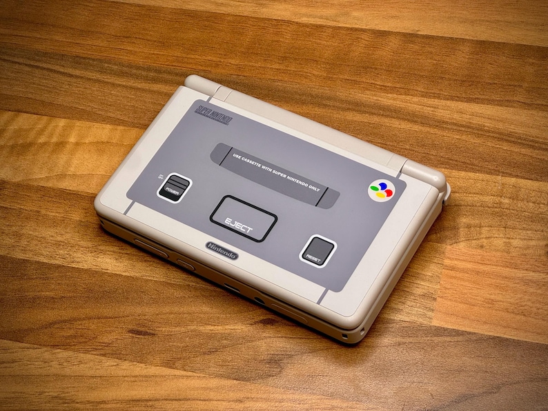 May include: A handheld Super Nintendo Entertainment System (SNES) in light grey and off-white. It has a grey central panel with an 'EJECT' button and a multi-coloured logo. The console is on a wooden surface.