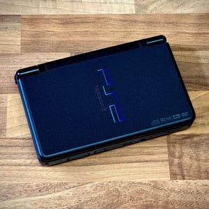 May include: A black PlayStation 2 console, featuring the PlayStation 2 logo in blue and green. The console is rectangular with rounded corners and is set against a wooden background.