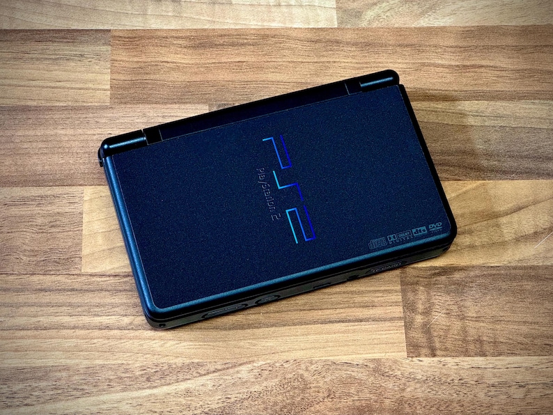 May include: A black PlayStation 2 console, featuring the PlayStation 2 logo in blue and green. The console is rectangular with rounded corners and is set against a wooden background.