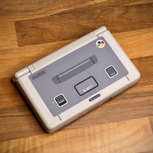 May include: A Super Nintendo handheld console in a light gray and off-white color scheme. The console features a gray center panel with the text "USE CASSETTE WITH SUPER NINTENDO ONLY" and an "EJECT" button. The Nintendo logo is visible.