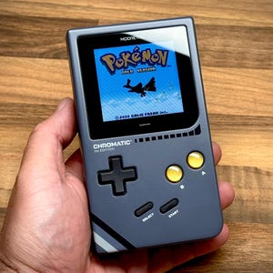 Puede incluir: Consola de juegos portátil gris que muestra el juego "Pokémon Gold Version". La consola tiene una cruceta negra, botones A y B amarillos y botones "SELECT" y "START". La pantalla muestra un cielo azul y una criatura voladora. El texto "CHROMATIC 16 EDITION" es visible.
