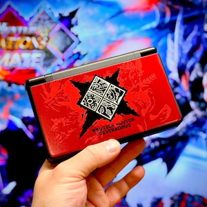May include: A red handheld games console with a black trim. The top cover displays a black and white graphic and the text "MONSTER HUNTER GENERATIONS ULTIMATE". The background is a blurred image of a video game.