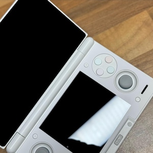 Pu&ograve; includere: Una console di gioco portatile bianca con due schermi, uno aperto e uno chiuso. La console ha un D-pad, pulsanti e un joystick. Il dispositivo &egrave; su una superficie di legno.