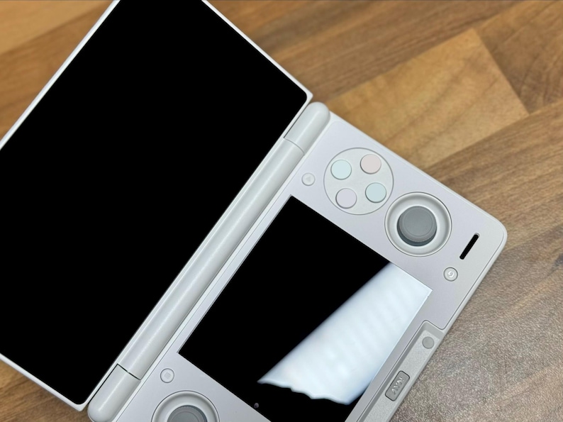 Pu&ograve; includere: Una console di gioco portatile bianca con due schermi, uno aperto e uno chiuso. La console ha un D-pad, pulsanti e un joystick. Il dispositivo &egrave; su una superficie di legno.