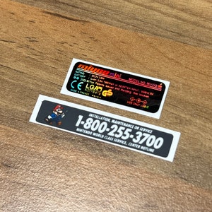 K&ouml;nnte beinhalten: Zwei schwarz-wei&szlig;e Aufkleber mit wei&szlig;em Text. Der obere Aufkleber ist ein schwarzes Rechteck mit wei&szlig;em Text, der "miyou mini MODEL NO MIYOO RATING DC5V 1.5A INPUT ADAPTER MUST USB-C 2022 MIYOO Pending See booklet CE LG4N GS USE ONLY USB-C" lautet. Der untere Aufkleber ist ein schwarzes Rechteck mit wei&szlig;em Text, der "INSTALLATION, MAINTENANCE OR SERVICE 1-800-255-3700 NINTENDO WORLD CLASS SERVICE, CENTER HOTLINE" lautet.