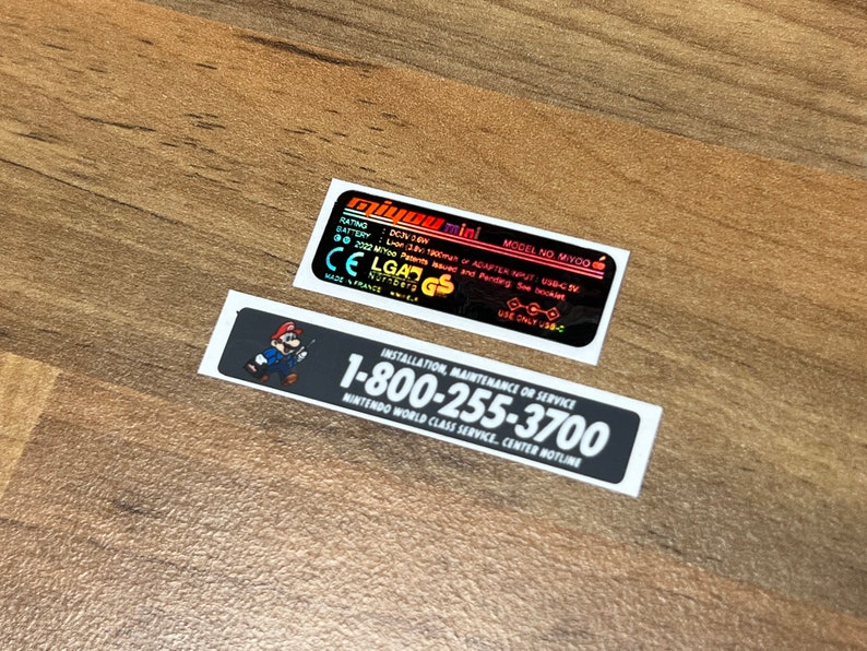 K&ouml;nnte beinhalten: Zwei schwarz-wei&szlig;e Aufkleber mit wei&szlig;em Text. Der obere Aufkleber ist ein schwarzes Rechteck mit wei&szlig;em Text, der "miyou mini MODEL NO MIYOO RATING DC5V 1.5A INPUT ADAPTER MUST USB-C 2022 MIYOO Pending See booklet CE LG4N GS USE ONLY USB-C" lautet. Der untere Aufkleber ist ein schwarzes Rechteck mit wei&szlig;em Text, der "INSTALLATION, MAINTENANCE OR SERVICE 1-800-255-3700 NINTENDO WORLD CLASS SERVICE, CENTER HOTLINE" lautet.