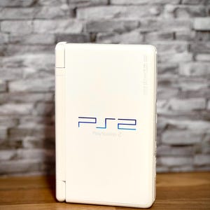 May include: A white PlayStation 2 console with the logo and text "PlayStation 2" in blue. The console is rectangular and has a hinged design. The background is a gray brick wall.