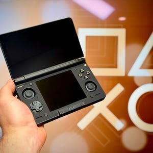 May include: Black handheld gaming console with two screens, buttons, and a directional pad. The console is held in a hand, with a blurred background featuring white geometric shapes. The console has the brand name "SONY" on it.