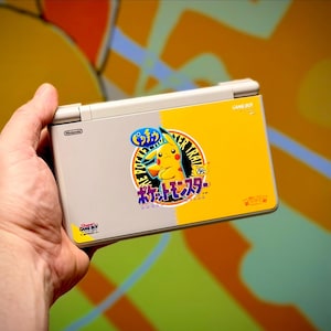May include: A Nintendo Game Boy Advance SP handheld console, featuring a two-tone design with a gray and yellow color scheme. The yellow side displays a Pikachu graphic with Japanese text. The console is held in a hand.