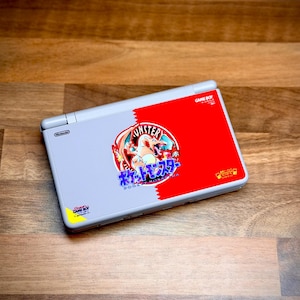 May include: A gray Nintendo DS Lite console with a red and white Pokemon-themed design. The design features the text "Monster" and Japanese characters. The console is on a wooden surface.