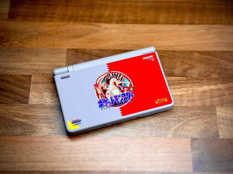 May include: A gray Nintendo DS Lite console with a red and white Pokemon-themed design. The design features the text "Monster" and Japanese characters. The console is on a wooden surface.
