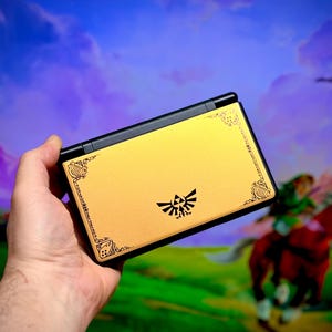 May include: A gold and black handheld gaming console featuring a decorative design and the Triforce symbol. The console has a gold finish with black accents and a detailed border. The background shows a scene from a video game.