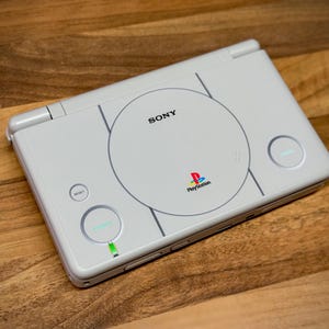 May include: A white handheld gaming console with the Sony logo and PlayStation branding. The console has a circular design on the top with a power button and a reset button. The console is on a wooden surface.