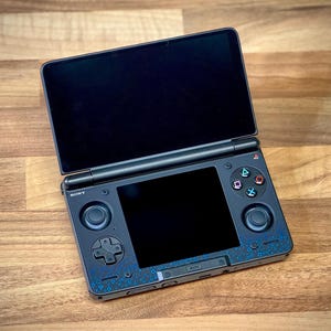 May include: A black Sony handheld gaming console with two screens, open on a wooden surface. The bottom screen has a black border with blue decorative patterns. The console features control buttons and a directional pad.