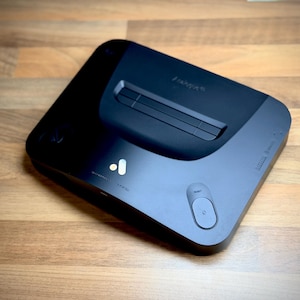 以下が含まれることがあります： 洗練されたモダンなデザインの黒いビデオゲーム機。コンソールは角が丸い長方形で、上部がわずかに盛り上がっています。電源ボタン、リセットボタン、ブランド名「freya」が特徴です。