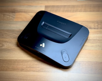 Puede incluir: Una consola de videojuegos negra con un diseño elegante y moderno. La consola tiene forma rectangular con esquinas redondeadas y una sección superior ligeramente elevada. Cuenta con un botón de encendido, un botón de reinicio y el nombre de la marca "freya".