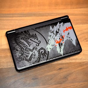 Puede incluir: Una consola de juegos portátil negra con un gráfico detallado en la cubierta superior. El diseño incluye un emblema de dragón tribal circular a la izquierda y una ilustración estilizada de una cabeza de criatura blanca y naranja a la derecha.