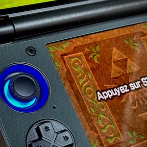 May include: Close-up of a black handheld gaming console with a blue illuminated control. The screen displays a brown and green design with the text "Appuyez sur START". A gold triangle logo is visible.